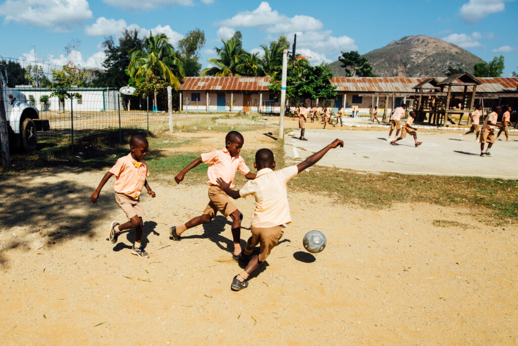 Three children are in the foreground playing football whilst in the background other children are also playing.