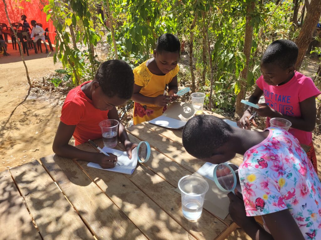 Four children are observing water with magnifying glasses. They are taking down notes in their books