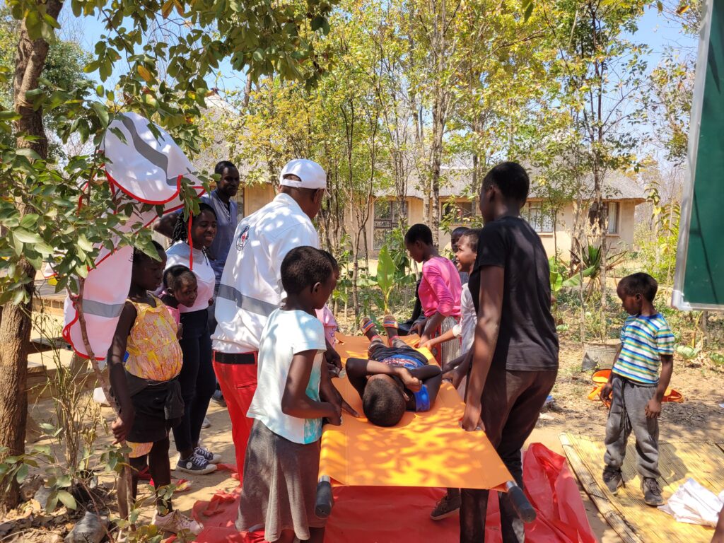 A child is laid on a stretcher whilst 5 other children and a first aid trainer are carrying the child. Other children are observing the exercise.